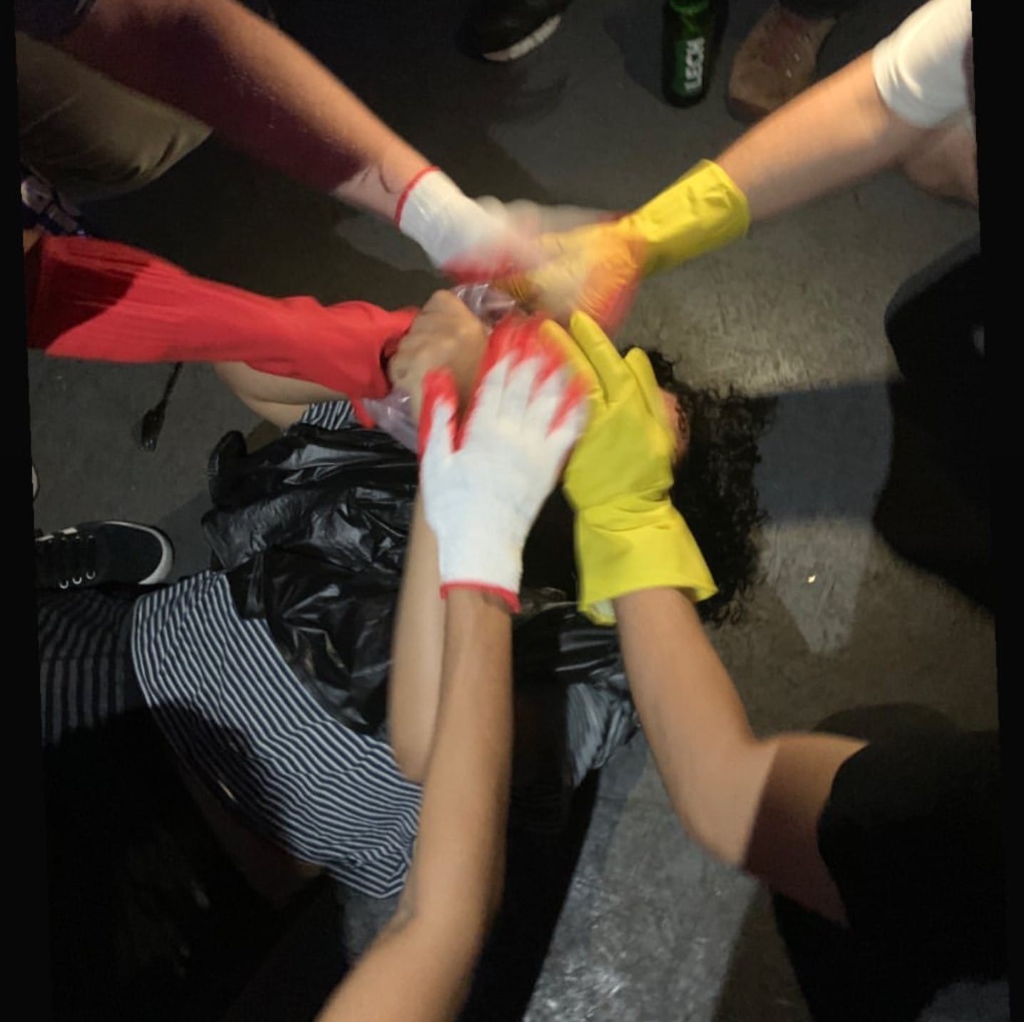 An overhead shot of a joining of five gloved hands which is shown above an covering the face of a person with curly hair in striped pajamas laying on a dark gray floor holding up a bag above their head.