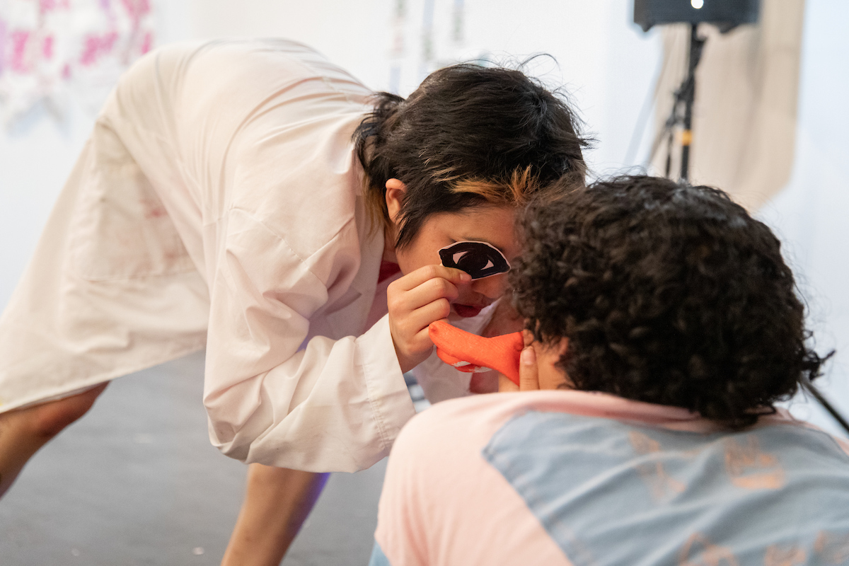 A shot of a small person in a white lab coat holding up a large cutout of an eye up to cover their one exposed eye. Covering the other half of their face is the back of a curlyhaired person holding up a glove to their mouth.
